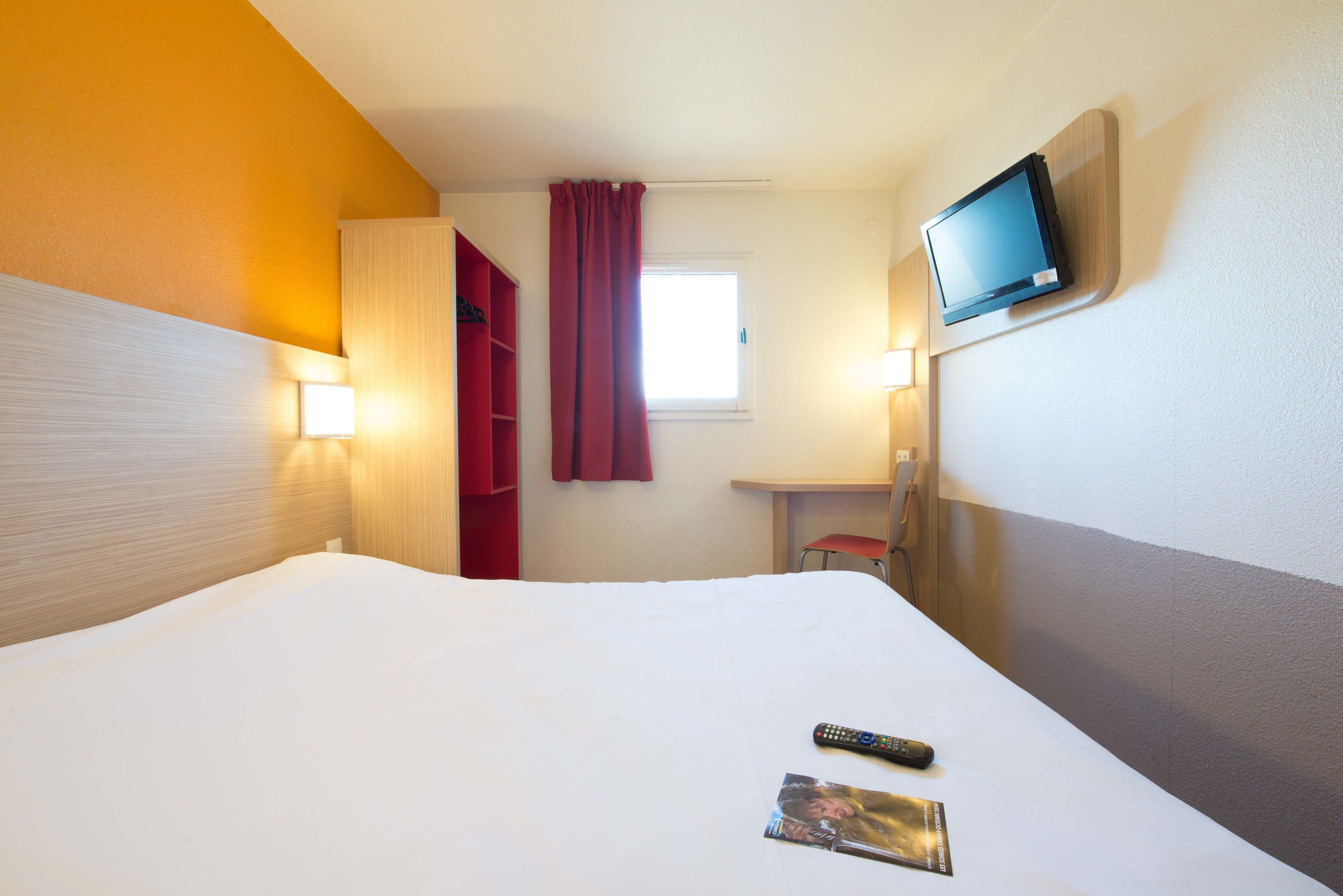A hotel room with a bed, a TV mounted on the wall, a window with curtains, and a bedside table.