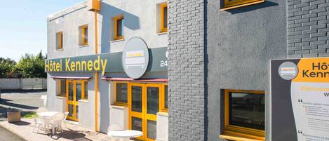 A hotel named "Hôtel Kennedy" with a modern facade, yellow-framed windows, and a signboard indicating room availability.