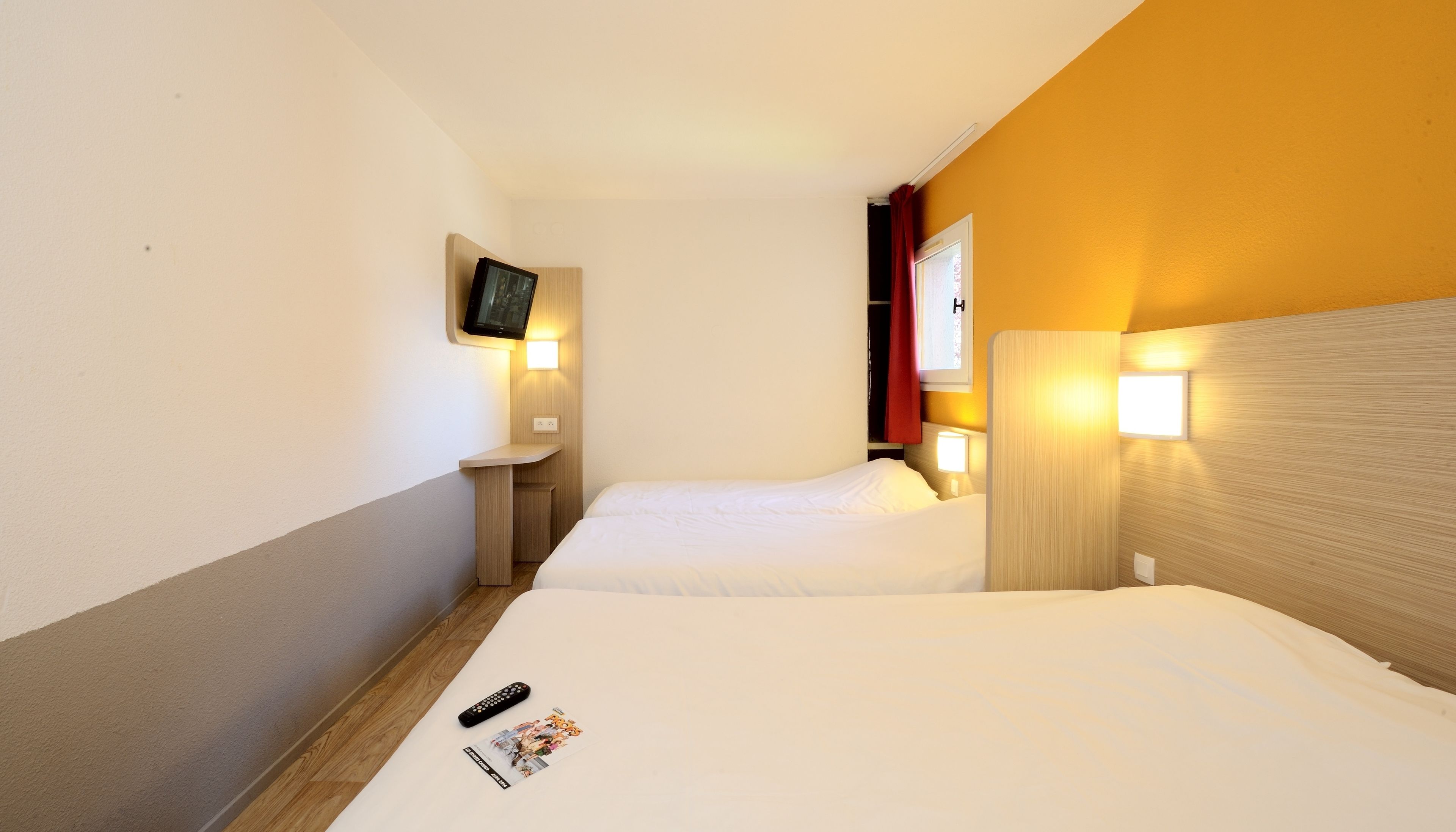 A hotel room with two beds, a TV mounted on the wall, bedside lamps, and a remote control on the floor.