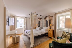 A bedroom with a four-poster bed, bedside tables, a green armchair, and a view of the street through large windows. - Boutiquehotel Amadeus (Salzburg)