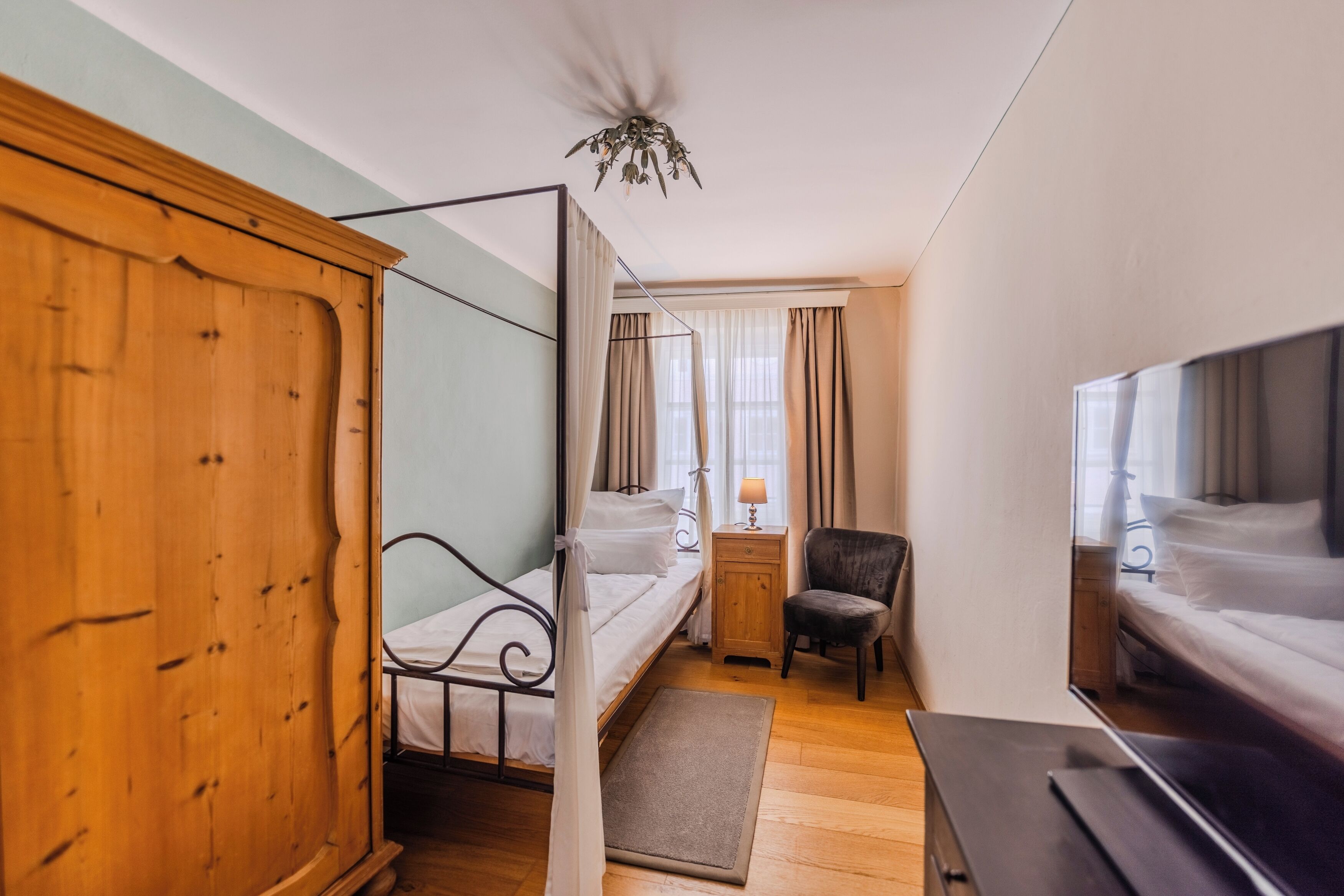 A bedroom with a wooden wardrobe, a single bed with a canopy, a desk with a chair, and a television.