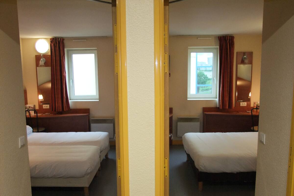 A mirrored hotel room with two beds, a window, a nightstand, and a lamp.