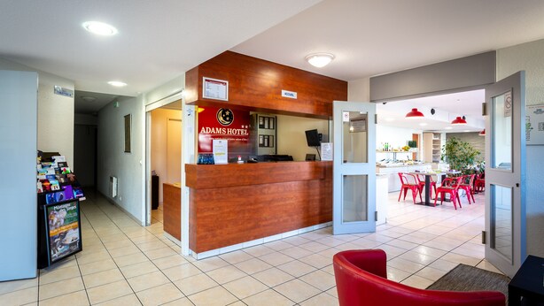 A hotel reception area with a wooden reception desk, a sign for Adams Hotel, and a dining area with red chairs and tables. - Adams Hotel (Metz)