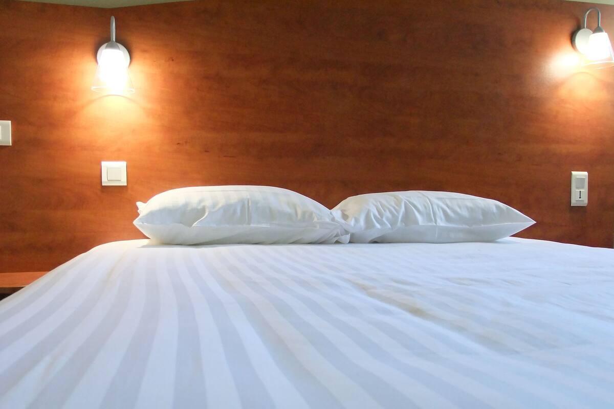 A neatly made bed with white linens and two pillows, a wooden headboard, and two wall-mounted light fixtures.