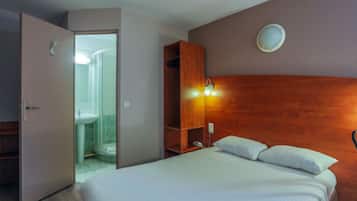 Habitación de hotel con cama, cabecera de madera, mesita de noche, estante y baño visible a través de una puerta abierta.
