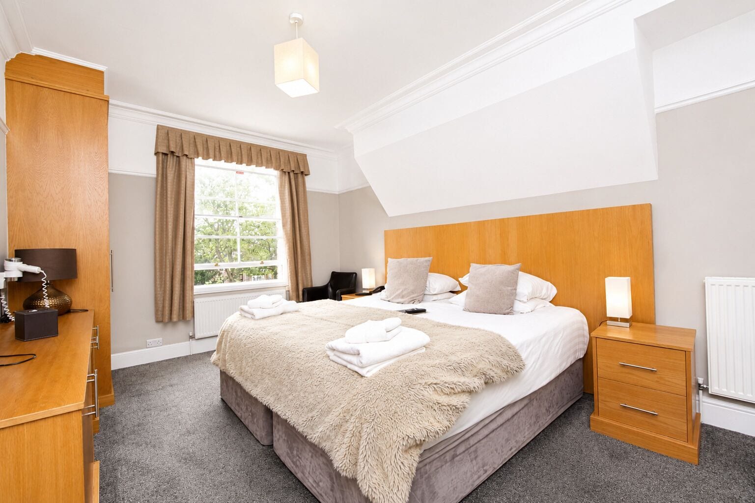 Quarto de hotel com uma cama grande, uma escrivaninha, uma cadeira, uma janela com cortinas e um radiador.