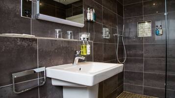 Un baño moderno con un lavamanos blanco, paredes de azulejos oscuros, un espejo y una zona de ducha.