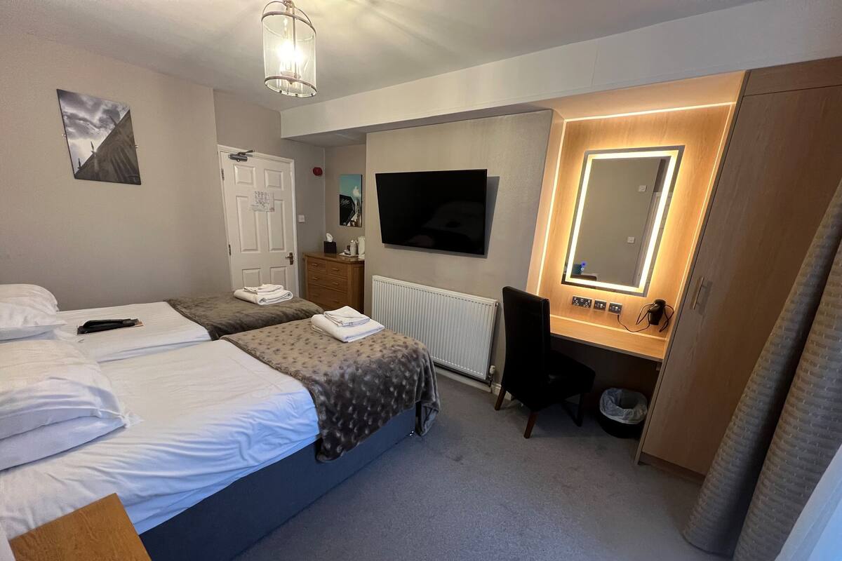 A hotel room with two beds, a desk with a chair, a TV, a mirror, and a radiator.
