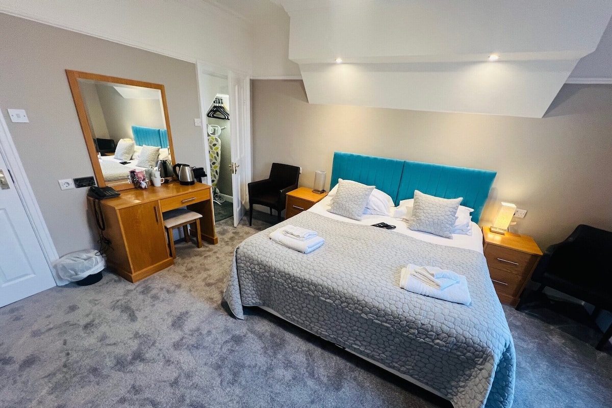 A hotel room with a large bed, a desk, a chair, a mirror, and a wardrobe.