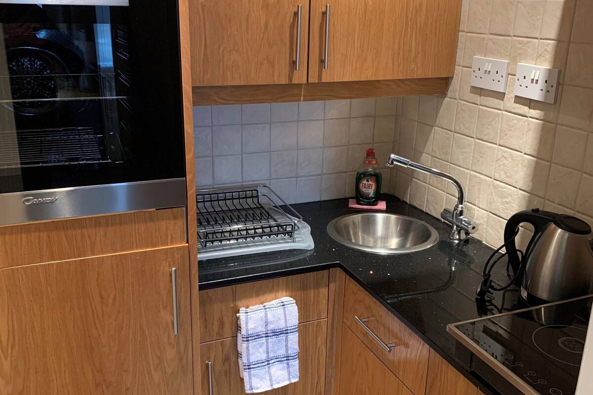A compact kitchen with wooden cabinets, a stainless steel range hood, a sink, and a kettle.