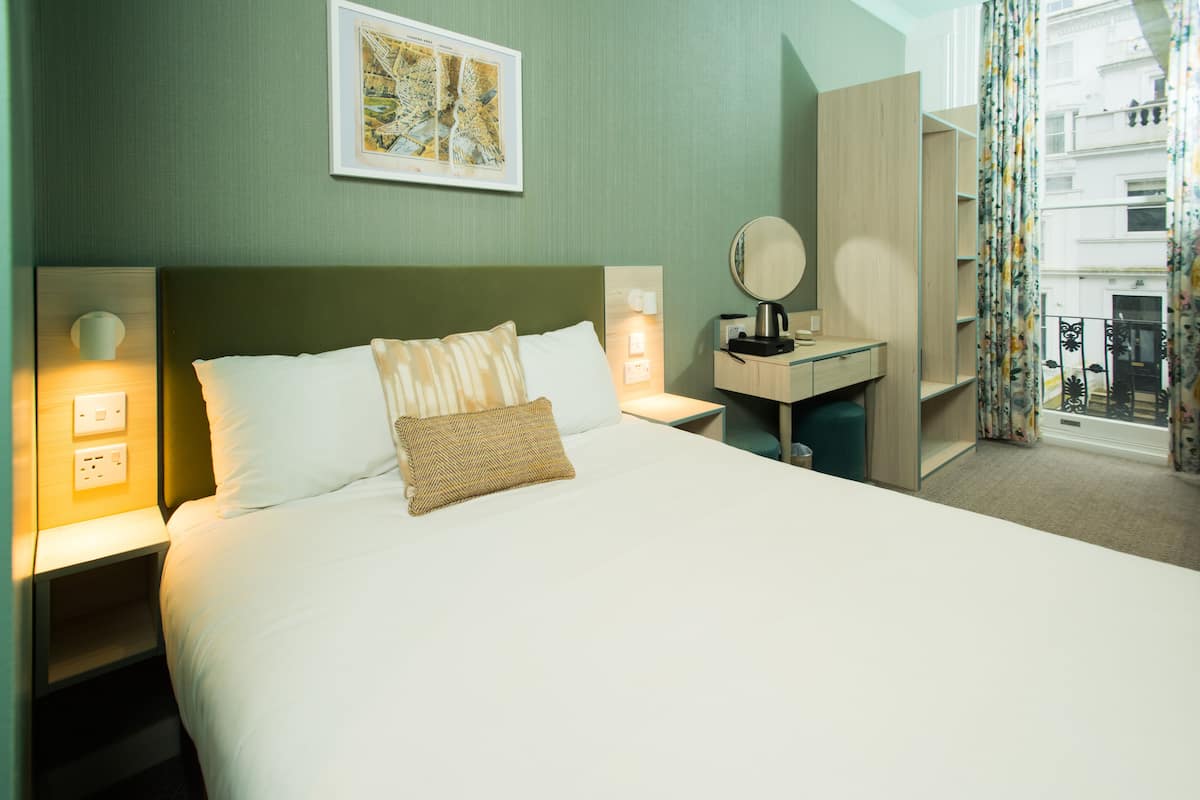 Un lit bien fait, avec du linge de lit blanc et un coussin décoratif marron, une table de chevet avec une lampe, un miroir et une petite commode en bois.
