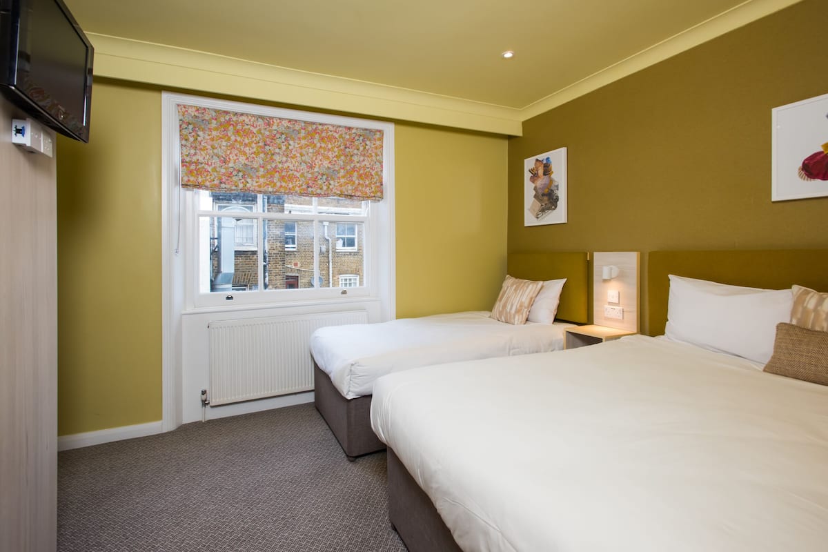 Une chambre d’hôtel avec deux lits, une fenêtre avec un store à motifs floraux, un radiateur et un téléviseur à écran plat fixé au mur.