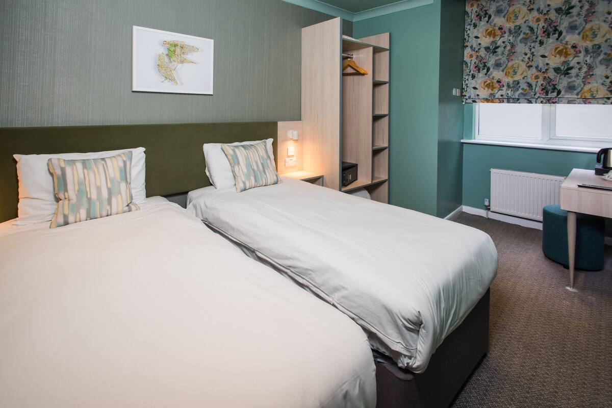 Une chambre d’hôtel avec deux lits, une armoire, un bureau et une fenêtre agrémentée de rideaux à motifs floraux.