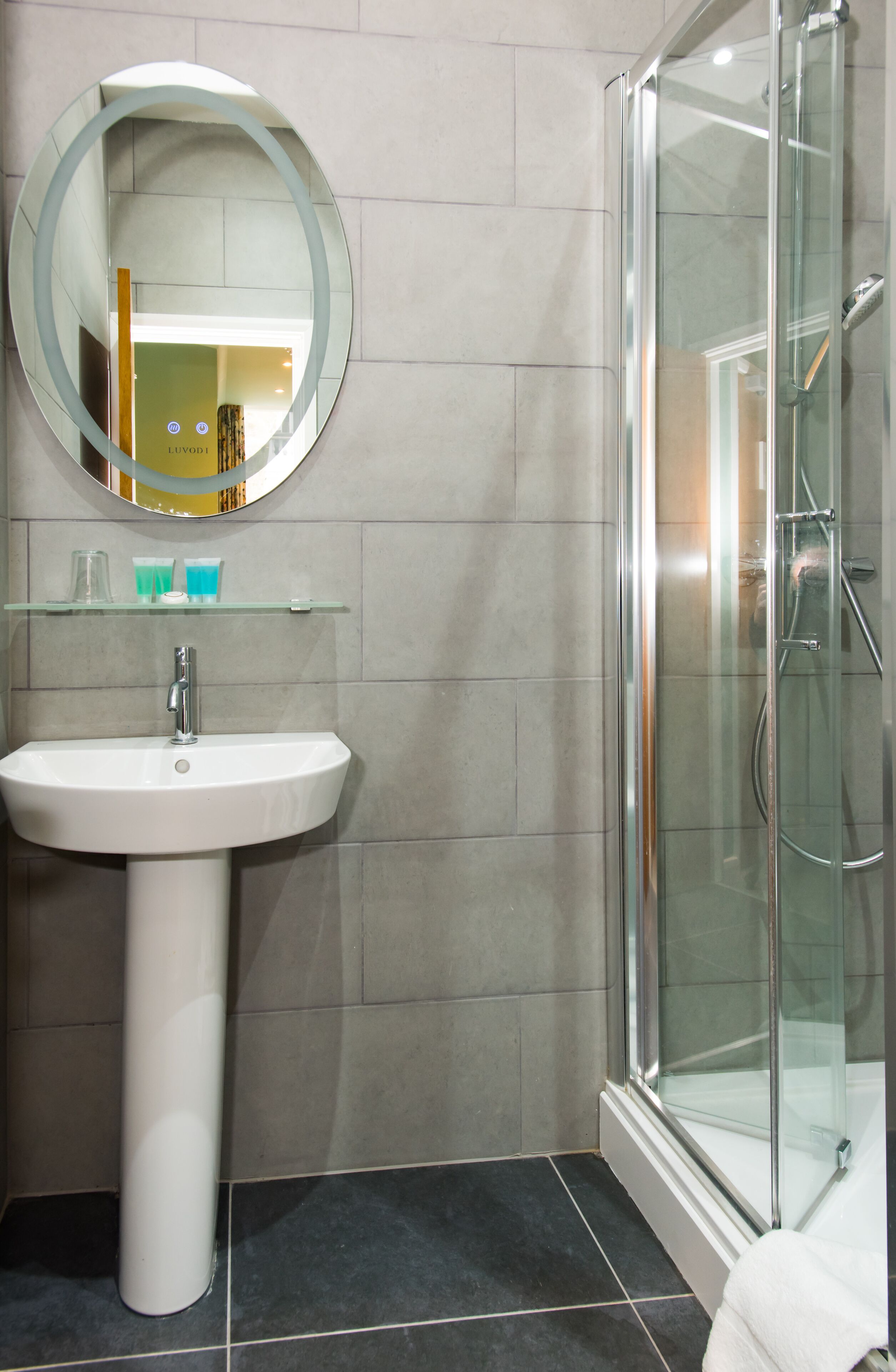 Ein Badezimmer mit einem weißen Waschbecken, einer Duschkabine aus Glas und einem runden Spiegel über dem Waschbecken.