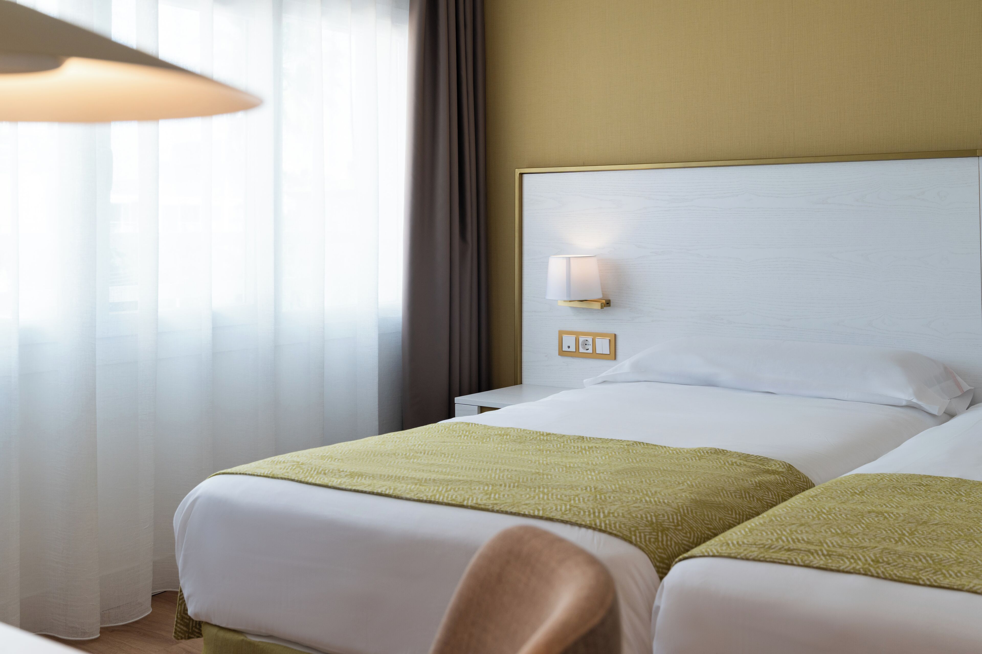 A hotel room with two beds, a nightstand, a lamp, and a window with curtains.