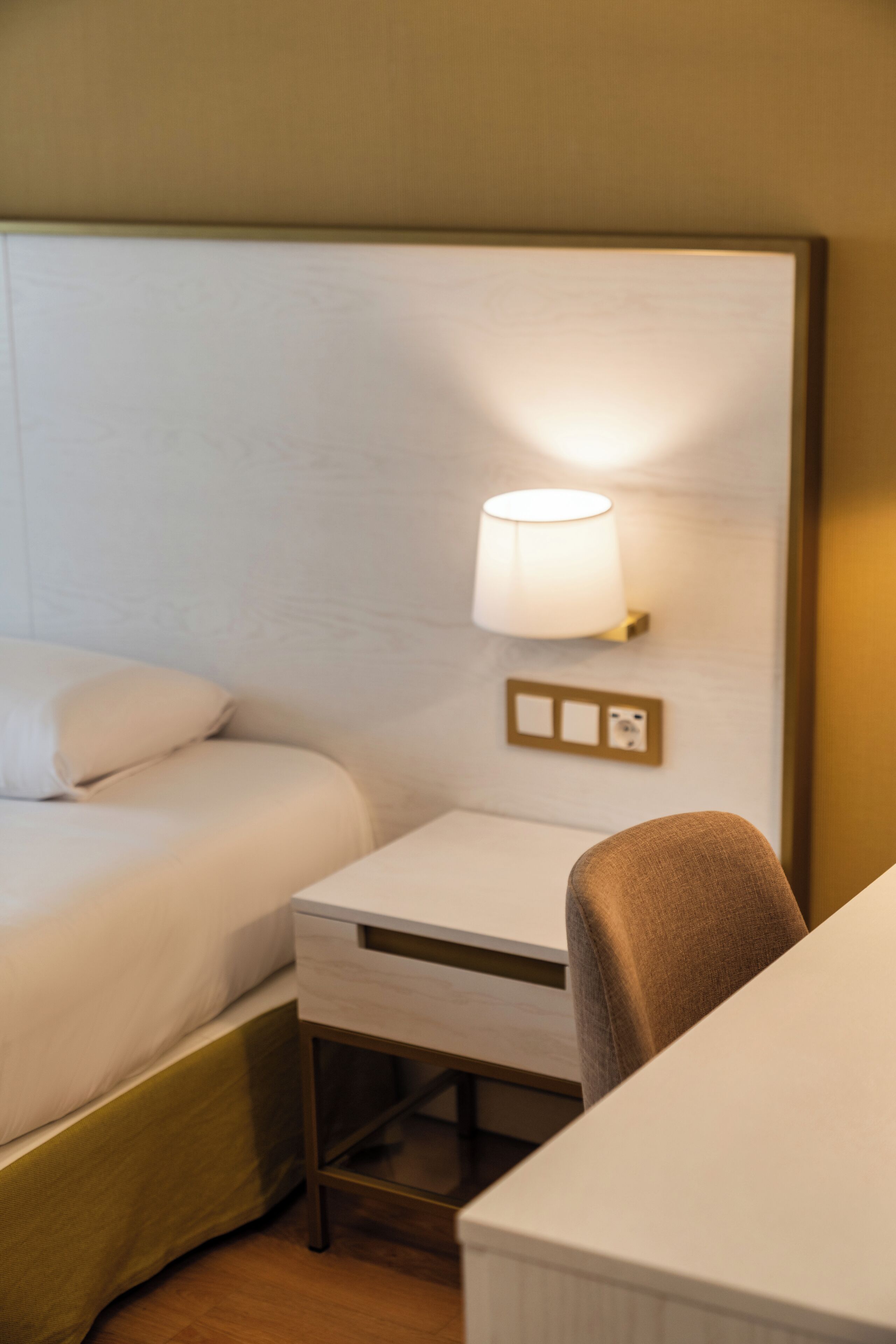 Habitación de hotel con una cama, un escritorio y una silla. Hay una lámpara en la pared sobre el escritorio.