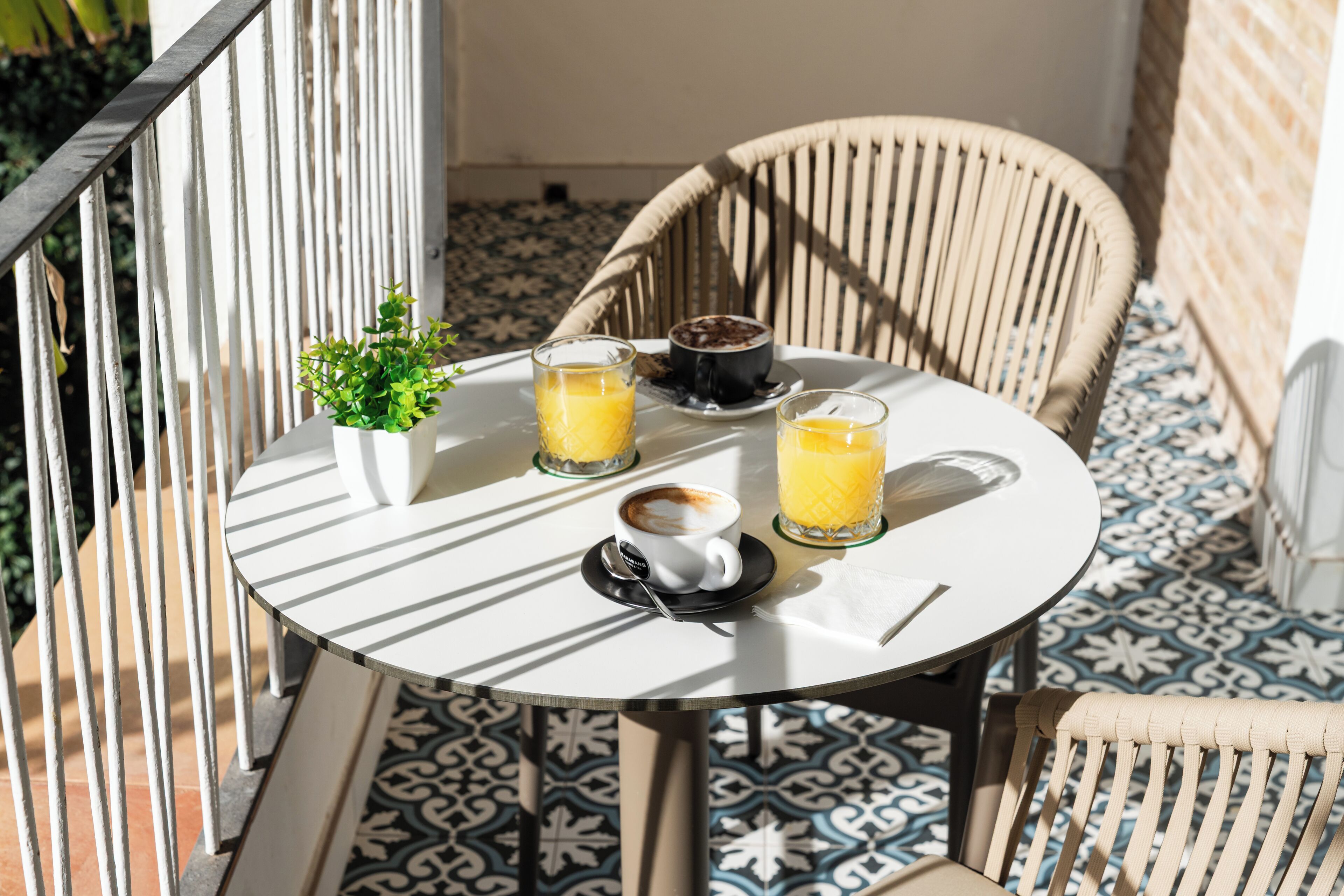 A round table with a white tablecloth, two glasses of orange juice, a cup of coffee, and a small potted plant on a balcony with patterned floor tiles and a wicker chair.