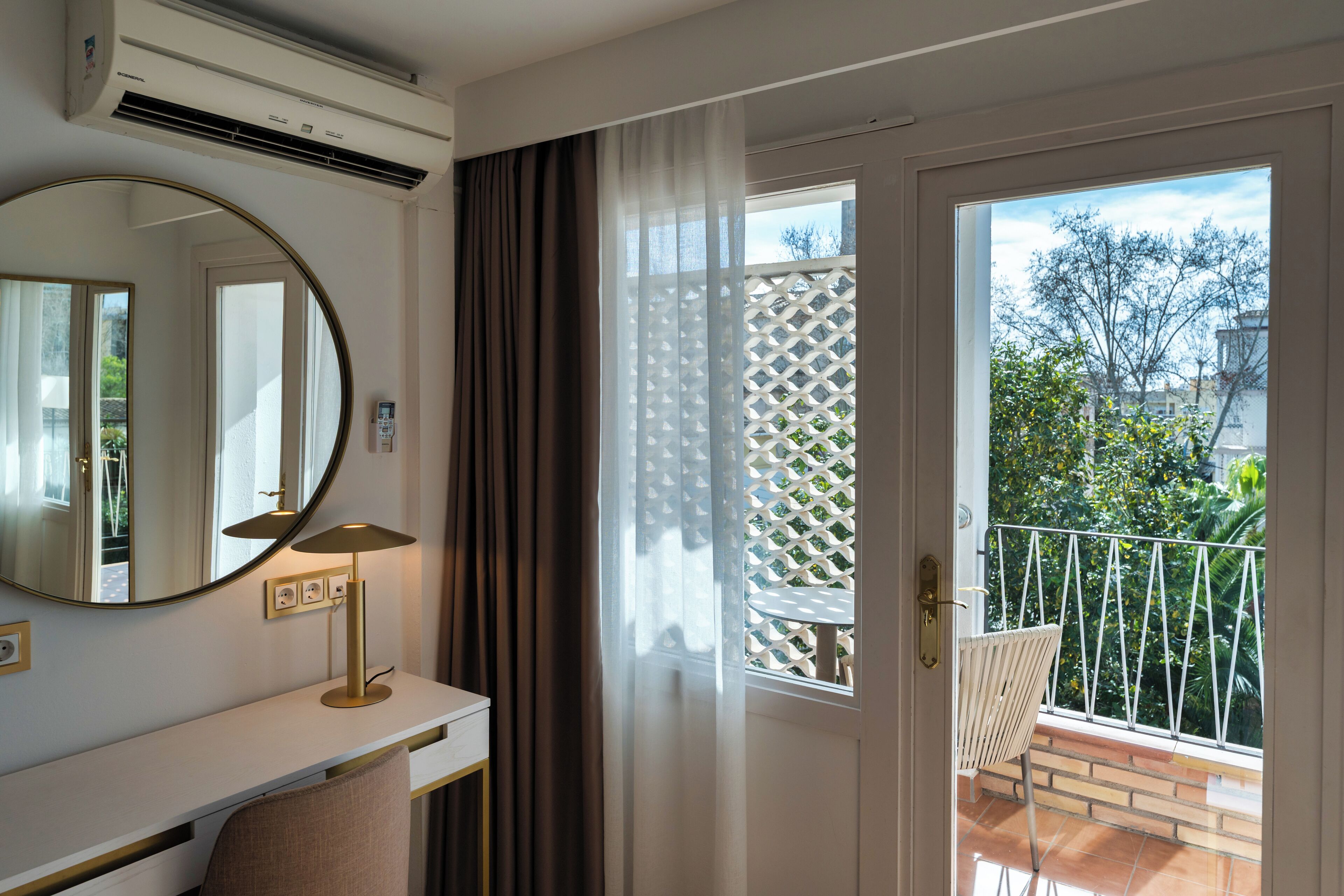 A room with a white desk, a round mirror, a lamp, and a view of a balcony with a white lattice fence and greenery.