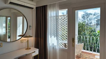 A room with a white desk, a round mirror, a lamp, and a view of a balcony with a white lattice fence and greenery.
