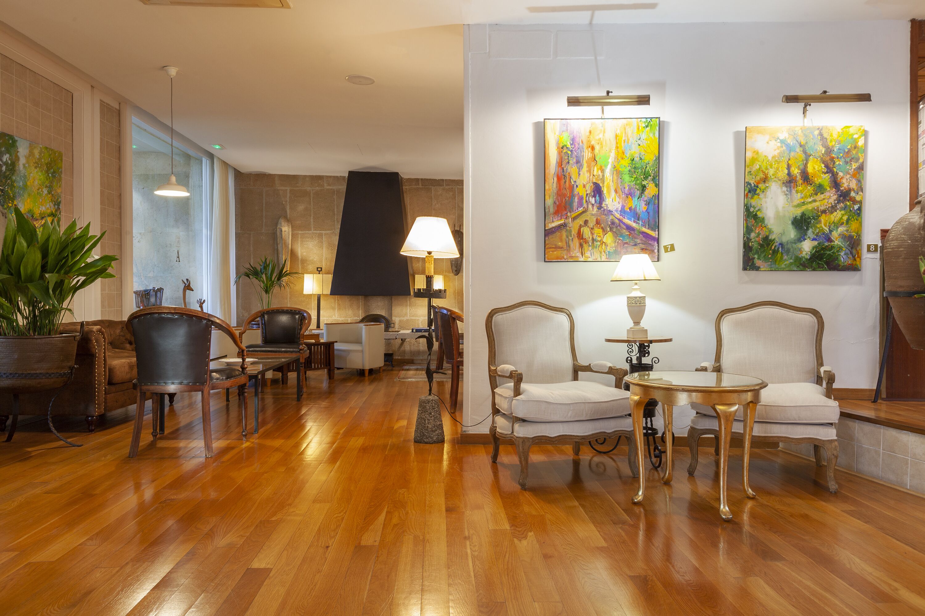 A spacious lobby with wooden flooring, leather armchairs, and a central table with a lamp. There are two paintings on the walls and potted plants.
