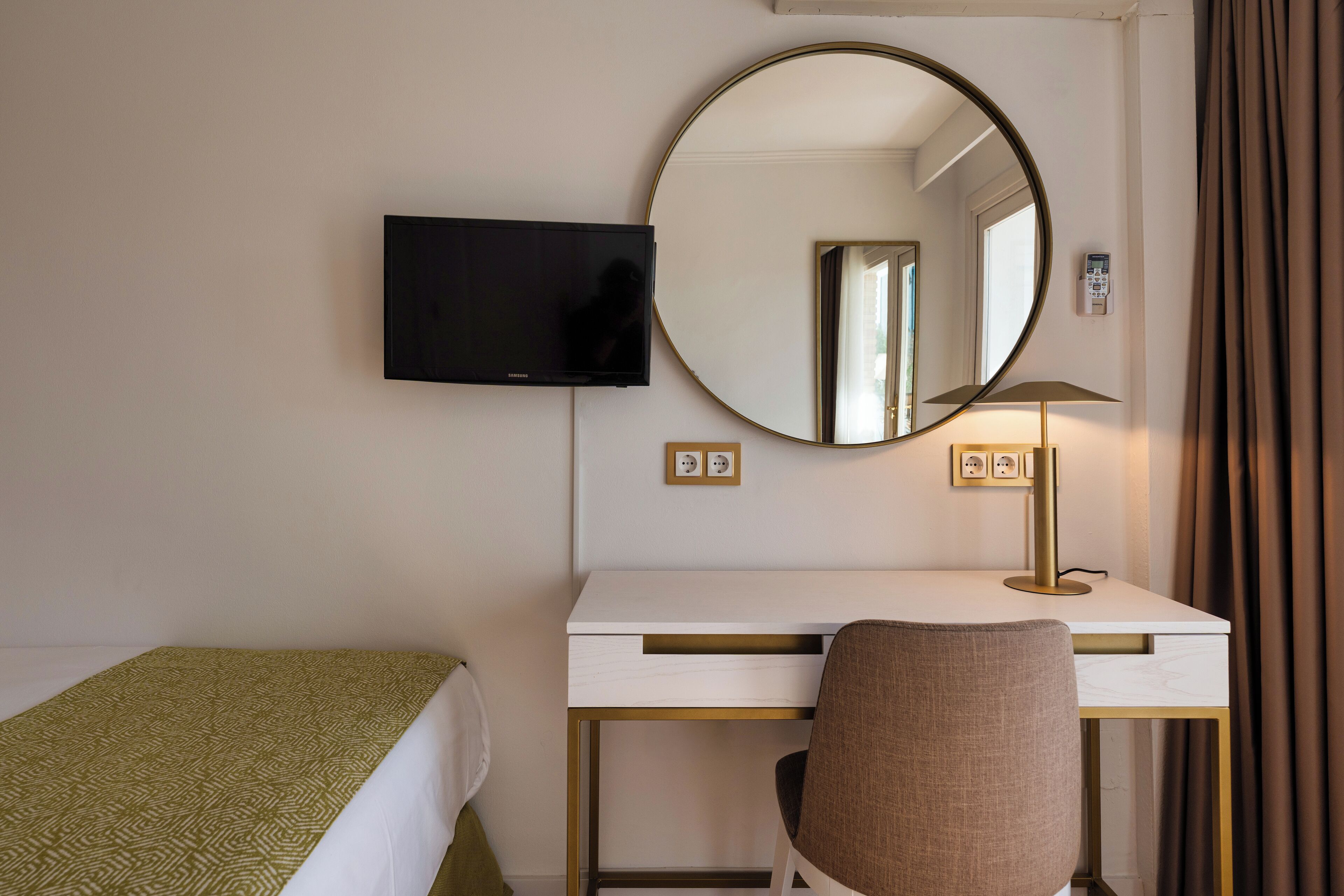 A hotel room with a bed, a desk with a computer, a chair, a TV, a round mirror, a lamp, and a window with curtains.
