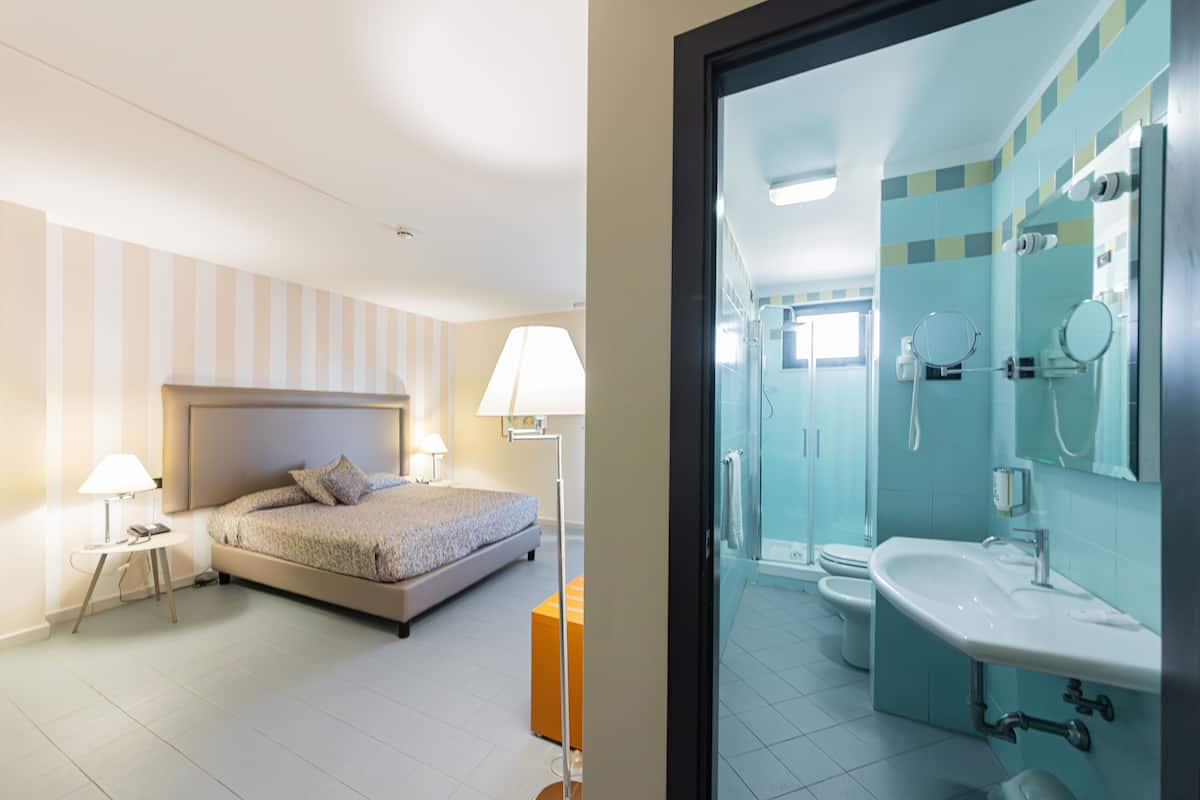 A hotel room with a bed, bedside table, lamp, and a bathroom with a sink and mirror.