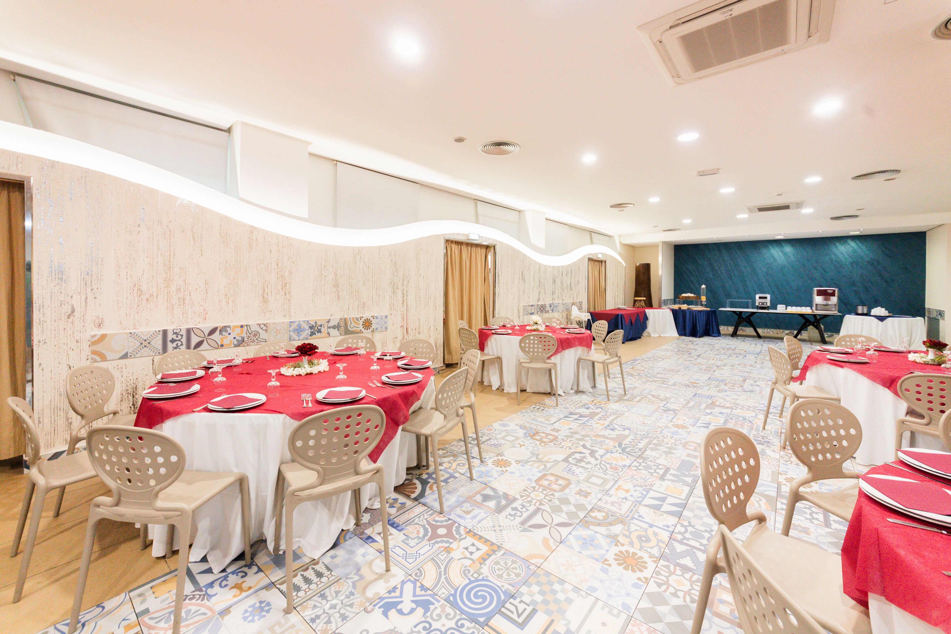 Un salón de banquetes con varias mesas dispuestas para comer, sillas color beige y un suelo de baldosas con diseño.