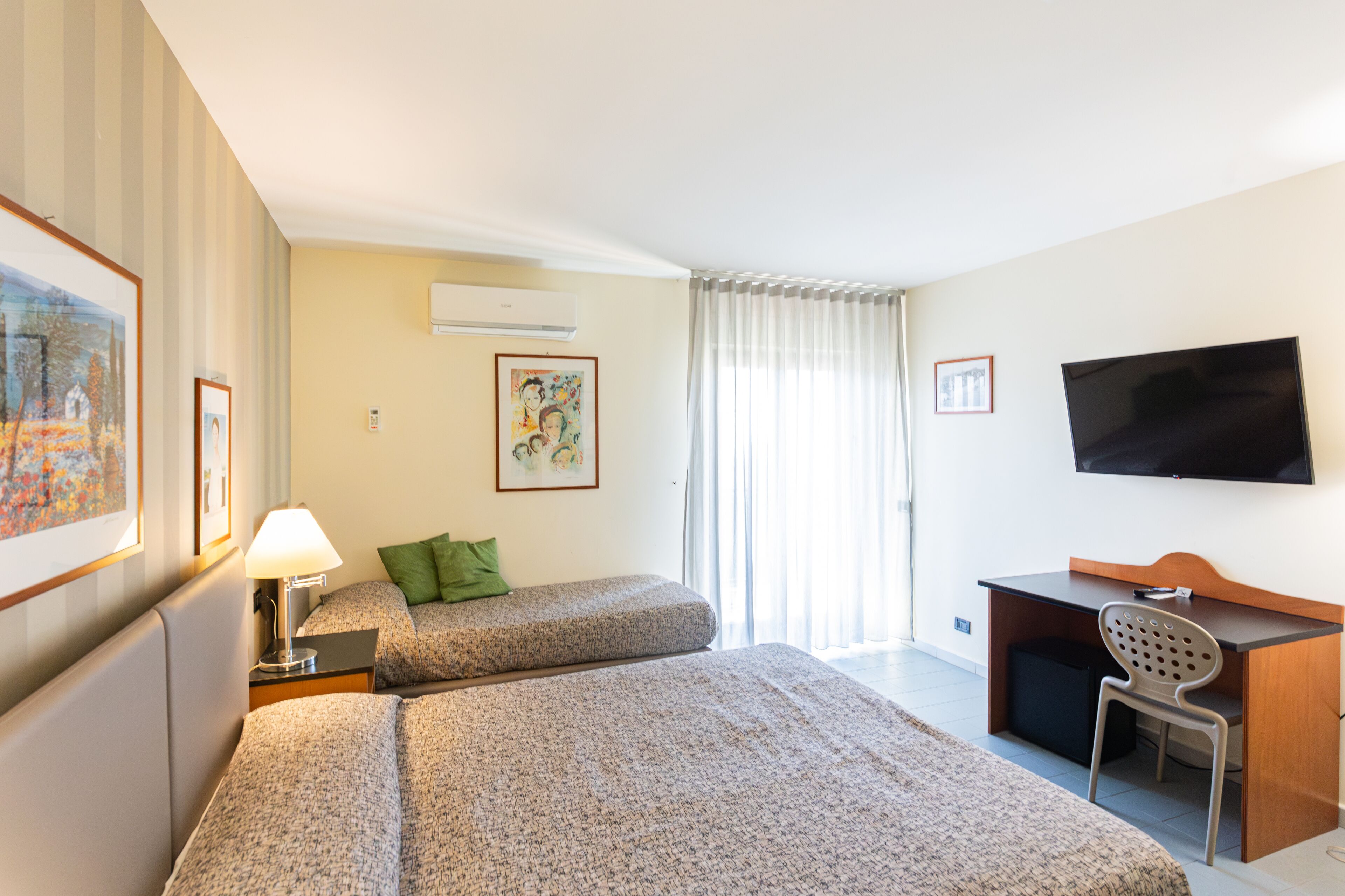 A hotel room with two beds, a desk, a chair, a TV, and artwork on the walls.