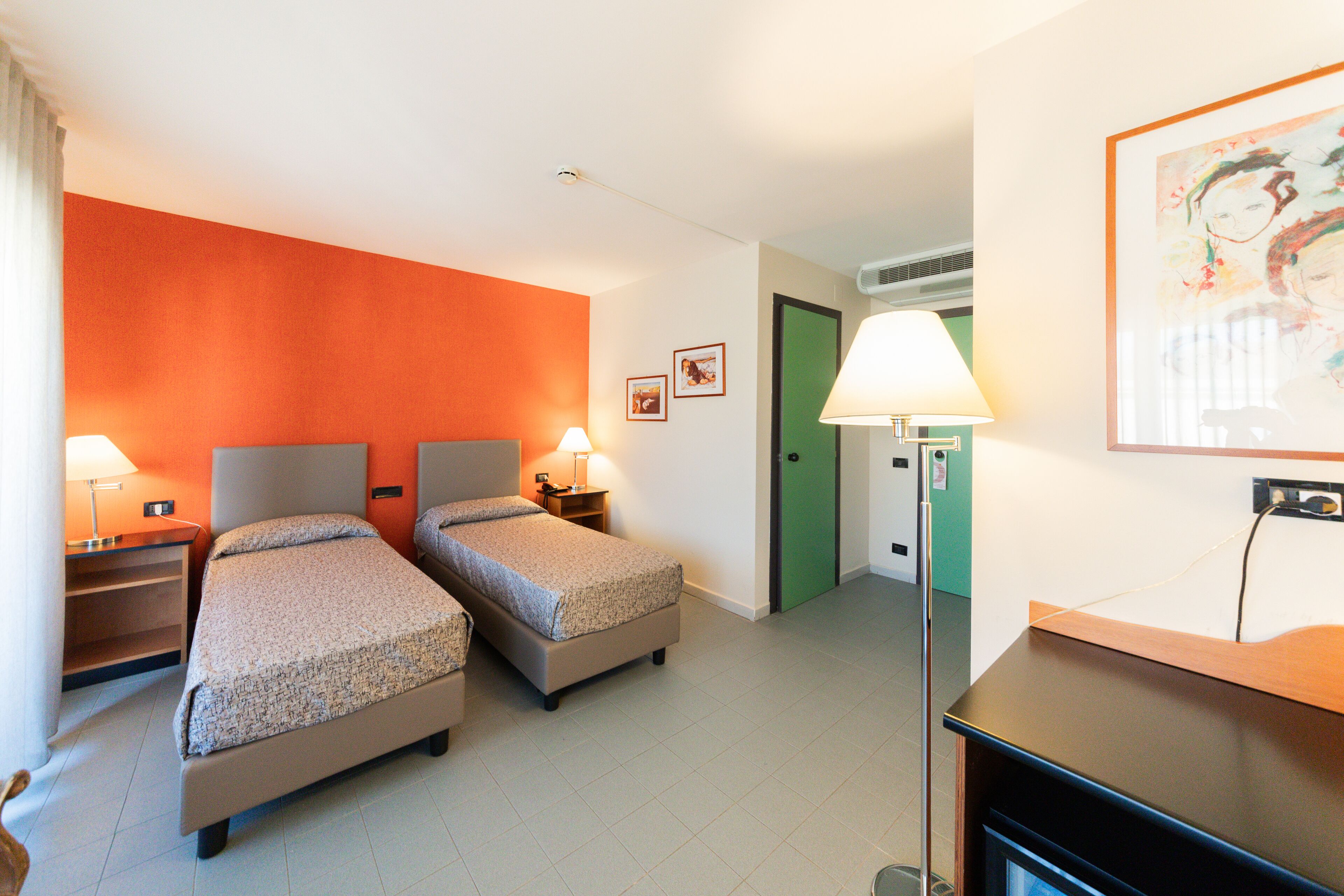 A hotel room with two beds, a desk, a chair, a lamp, and a painting on the wall.