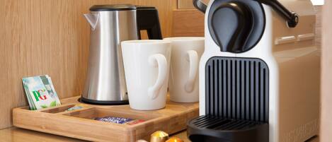 Una cafetera, dos tazas blancas, un hervidor de acero inoxidable y una bandeja de madera con cápsulas de café y una revista.