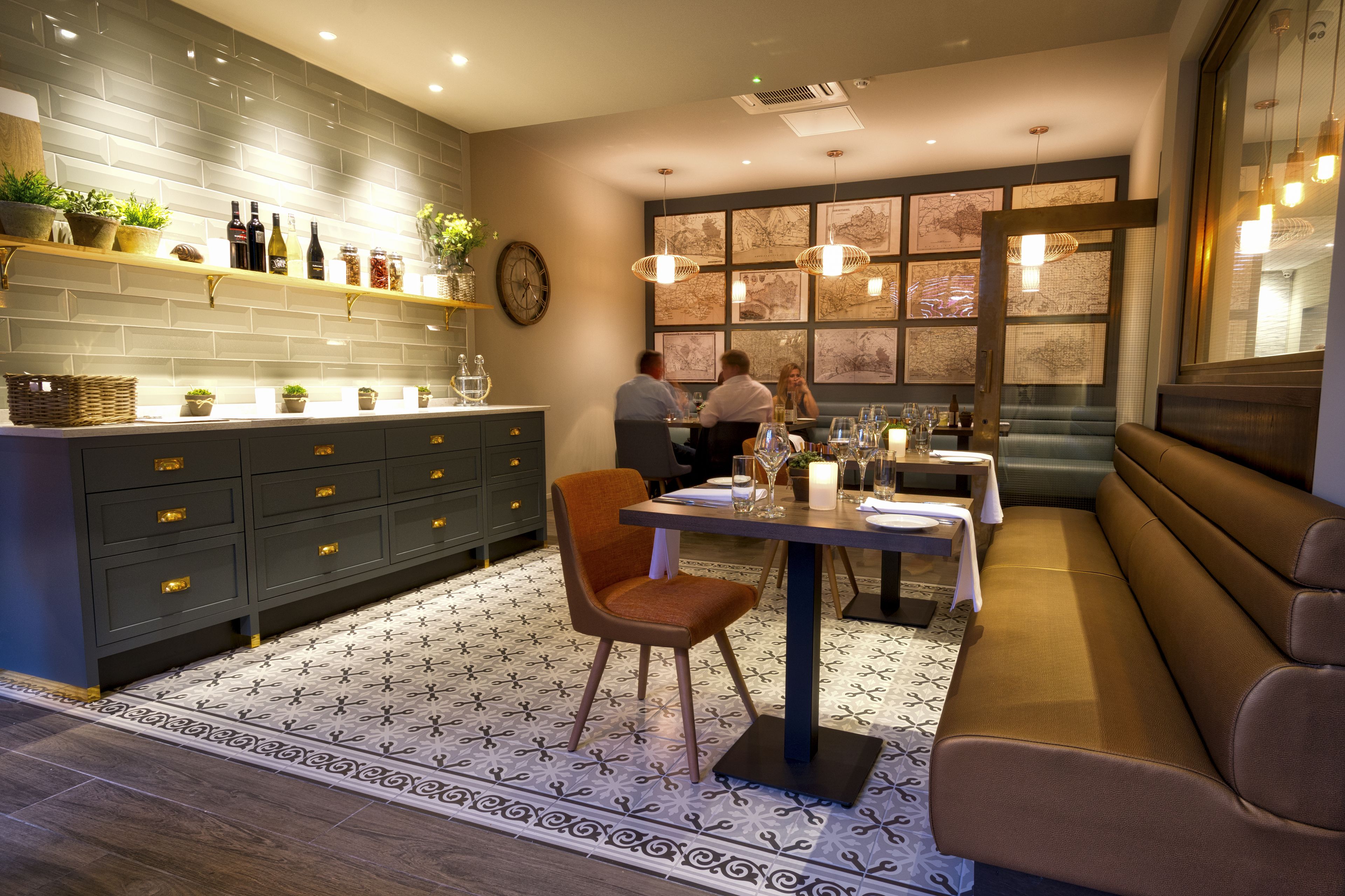 A modern restaurant interior with a dark grey tiled wall, wooden shelves with plants and bottles, a patterned floor, and a seating area with a bench and table.