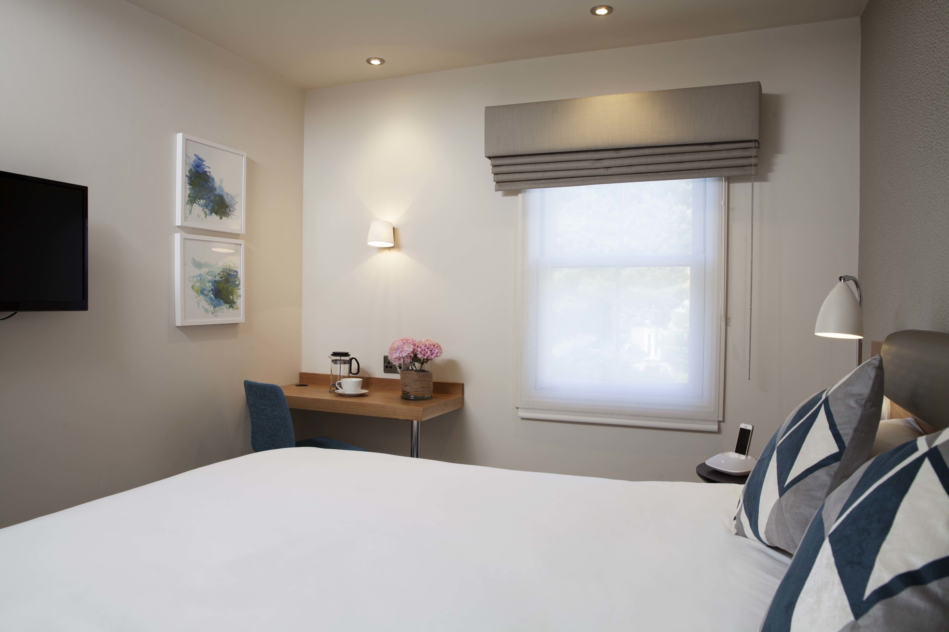 A hotel room with a bed, a desk with a chair, a TV mounted on the wall, and a window with a blind.