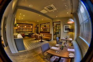 A modern dining area with a round table set for two, a sofa, and a kitchen in the background. - The Orchid Hotel (Bournemouth)