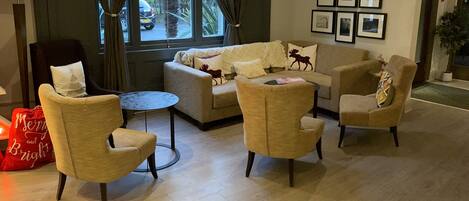 A modern living room with a sofa, two armchairs, a coffee table, and a lamp. There are curtains on the windows, and several framed pictures on the wall.
