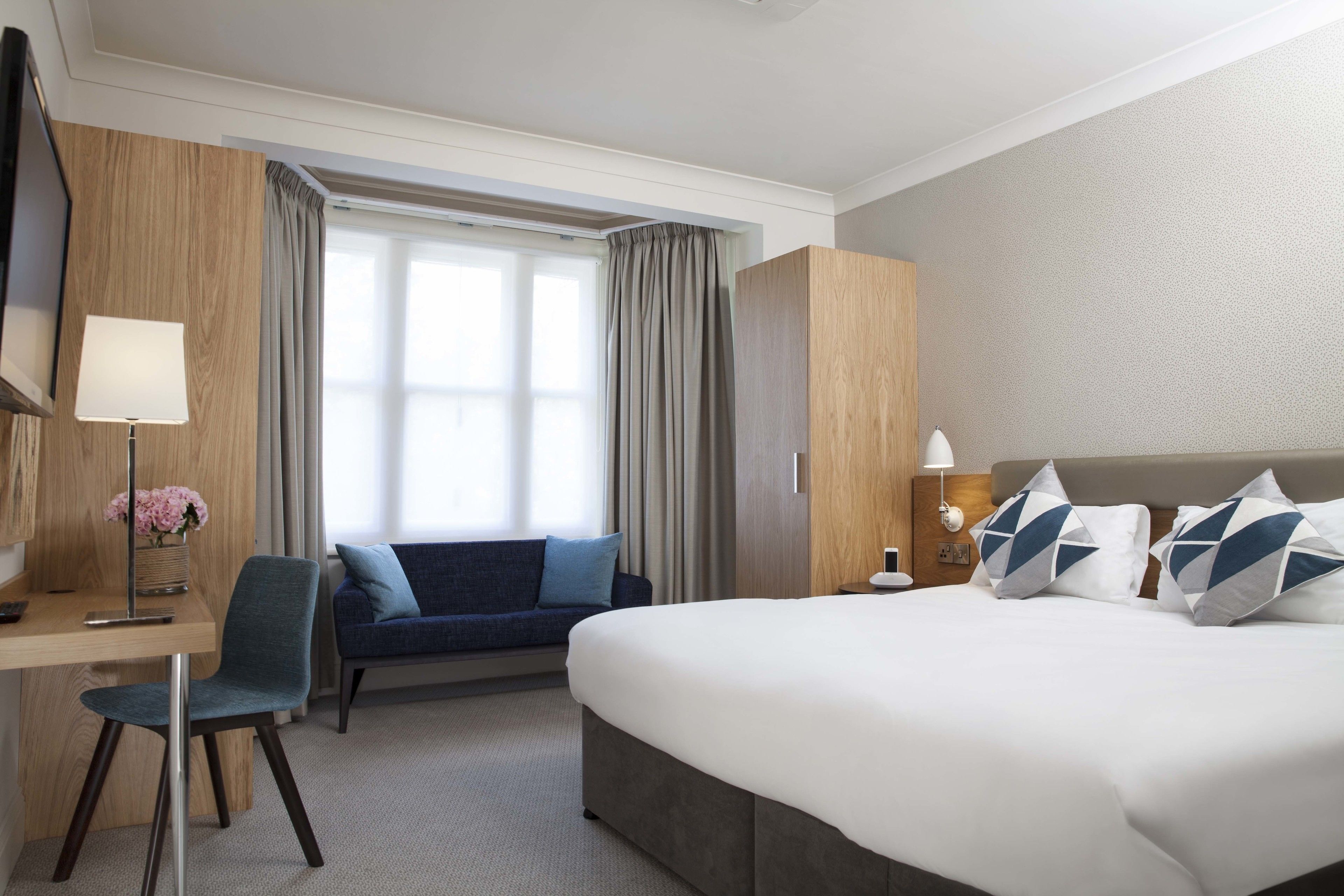 A hotel room with a bed, a desk, a chair, a sofa, a TV, and a window with curtains.