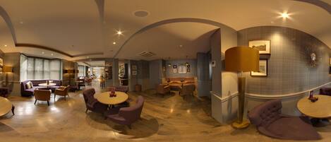 Eine geräumige Lounge mit runden Tischen und violetten Sitzgelegenheiten, einer Bar und einer großen Stehlampe.