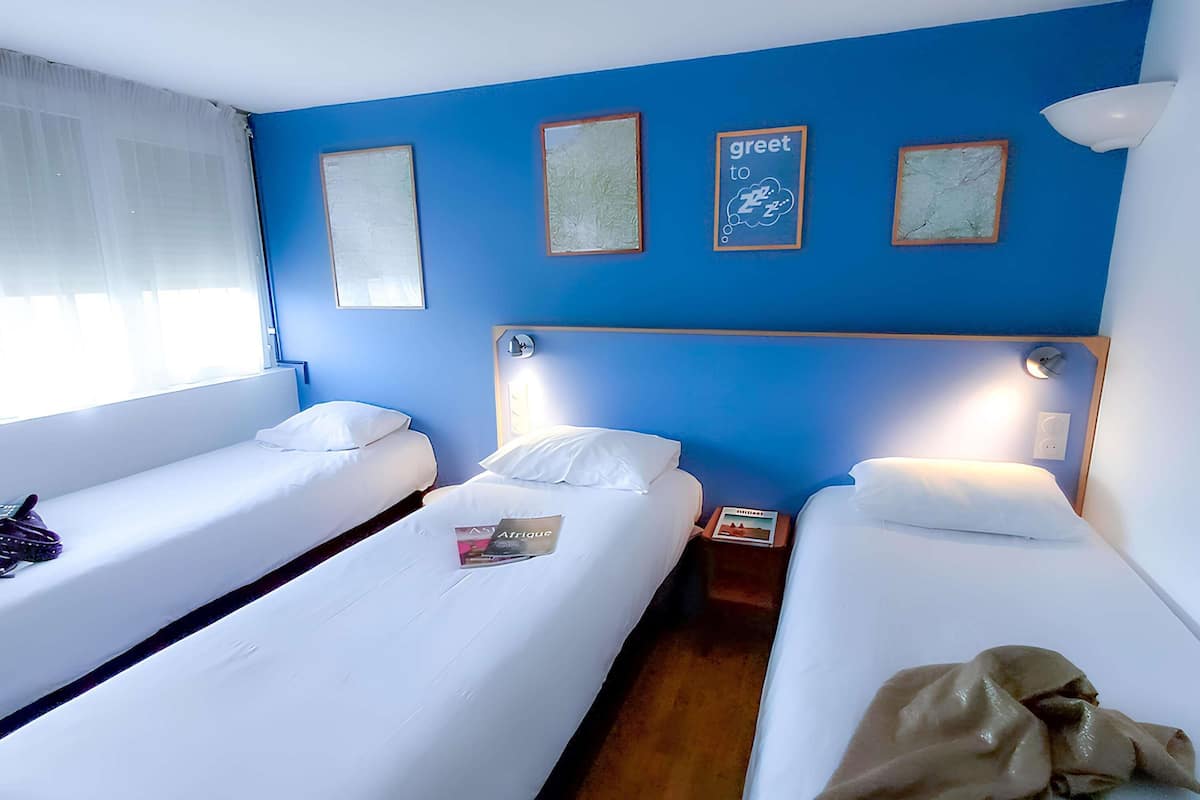 A hotel room with two single beds, a nightstand with a lamp, and a window with sheer curtains.