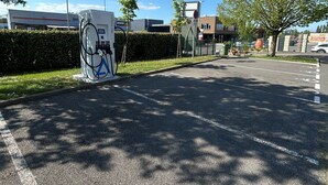 A parking lot with a fuel pump, a hedge, and a building in the background.