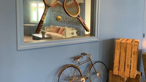 A room with a large mirror reflecting tennis rackets and a bicycle, a wooden crate, and a blue wall.
