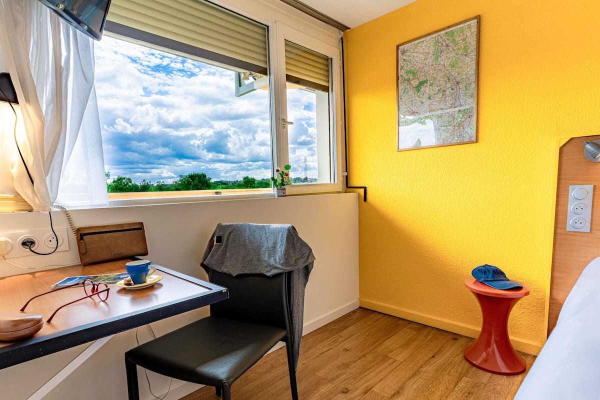 A room with a wooden floor, a desk with a chair, a window with blinds, a map on the wall, and a small red table with a cap on it.