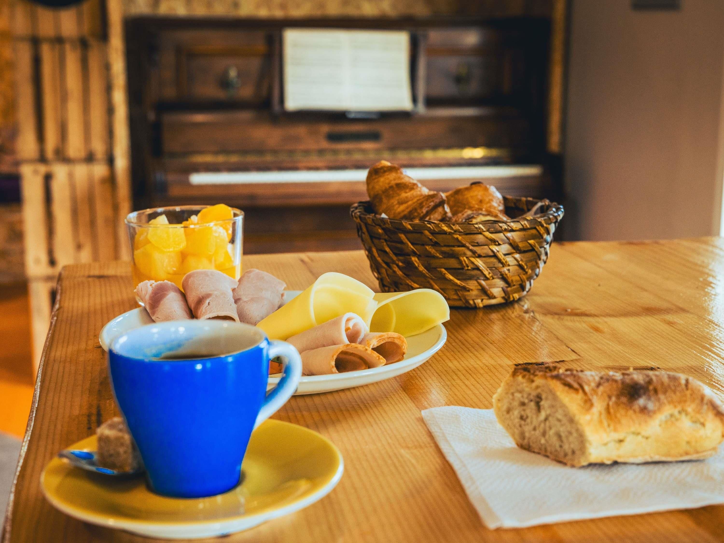 Un desayuno con una cesta de pan, un plato de queso y jamón, un vaso de zumo de naranja y una taza azul sobre una mesa de madera.