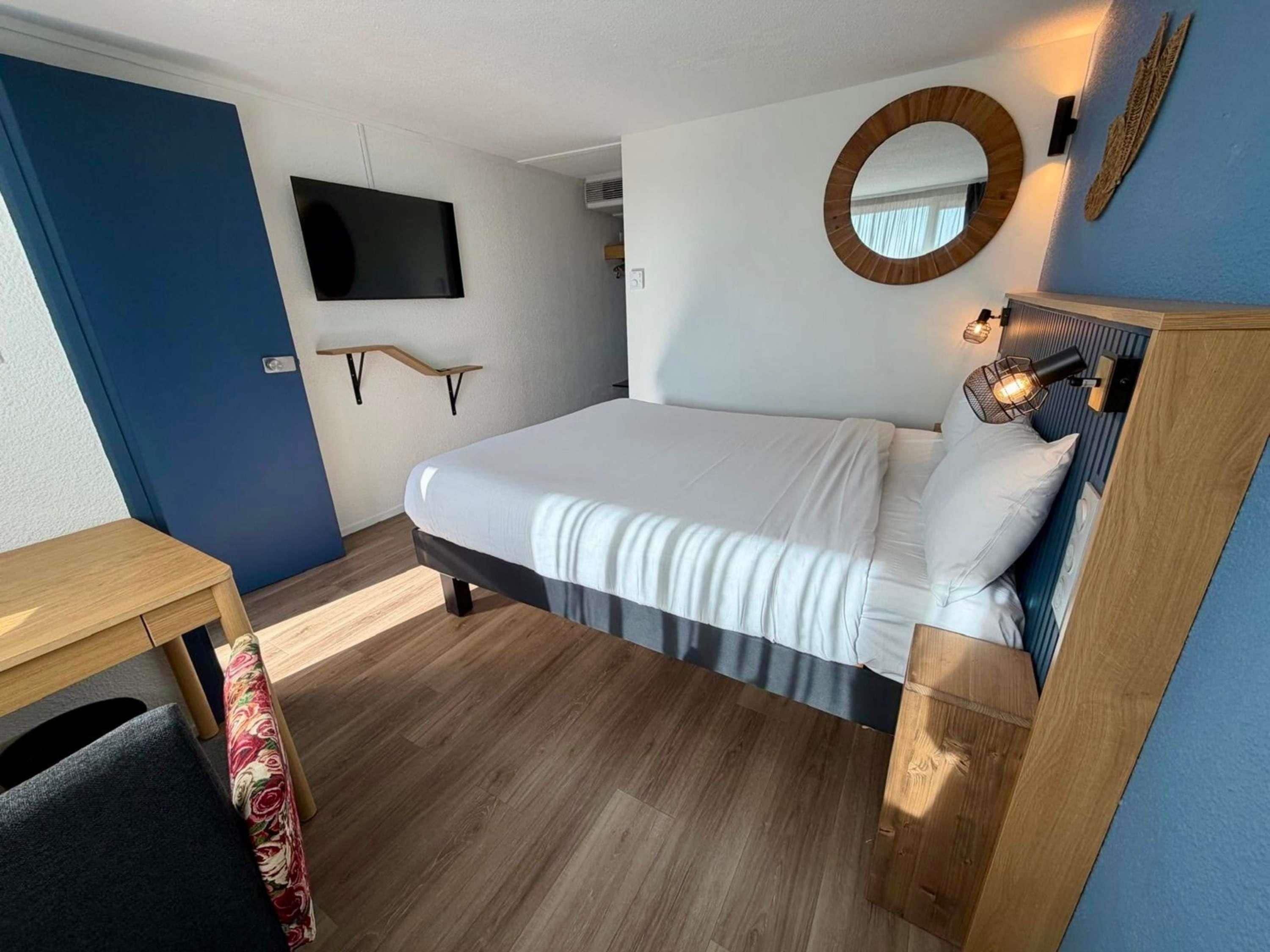 A hotel room with a bed, a television, a wall-mounted mirror, and a bedside table.