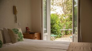 A bedroom with a bed, a bedside table, a telephone, and a window with a view of a garden and outdoor seating. - Hôtel Du Soleil (Saint-Raphael)
