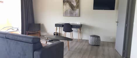Una sala de estar moderna con un sofá gris, una mesita, un escritorio con dos sillas, un televisor de pared y un otoman con estampado.