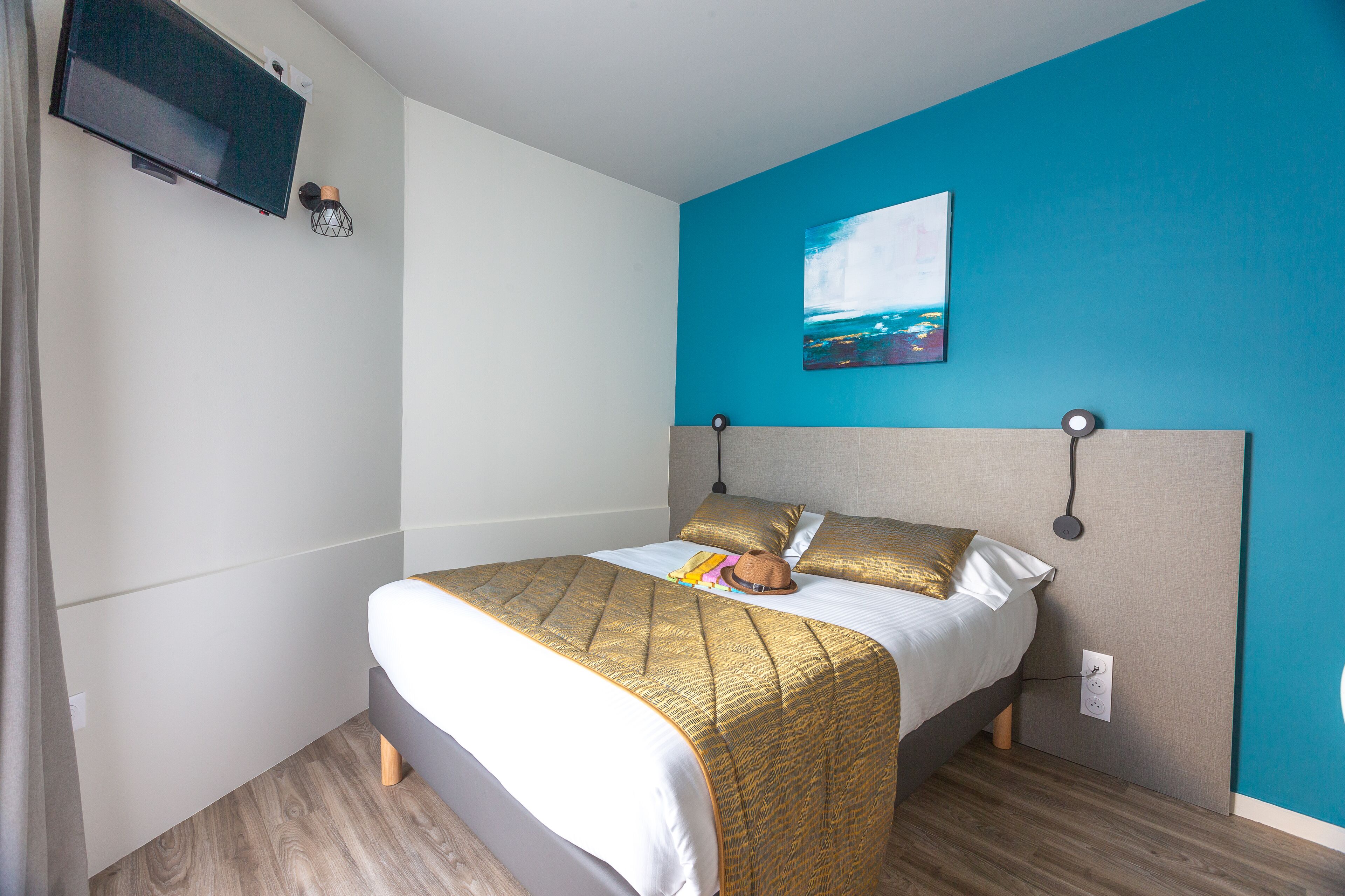 Una cama bien hecha con una colcha dorada y blanca, un televisor de pantalla plana montado en la pared, y un cuadro de un paisaje en la pared azul.