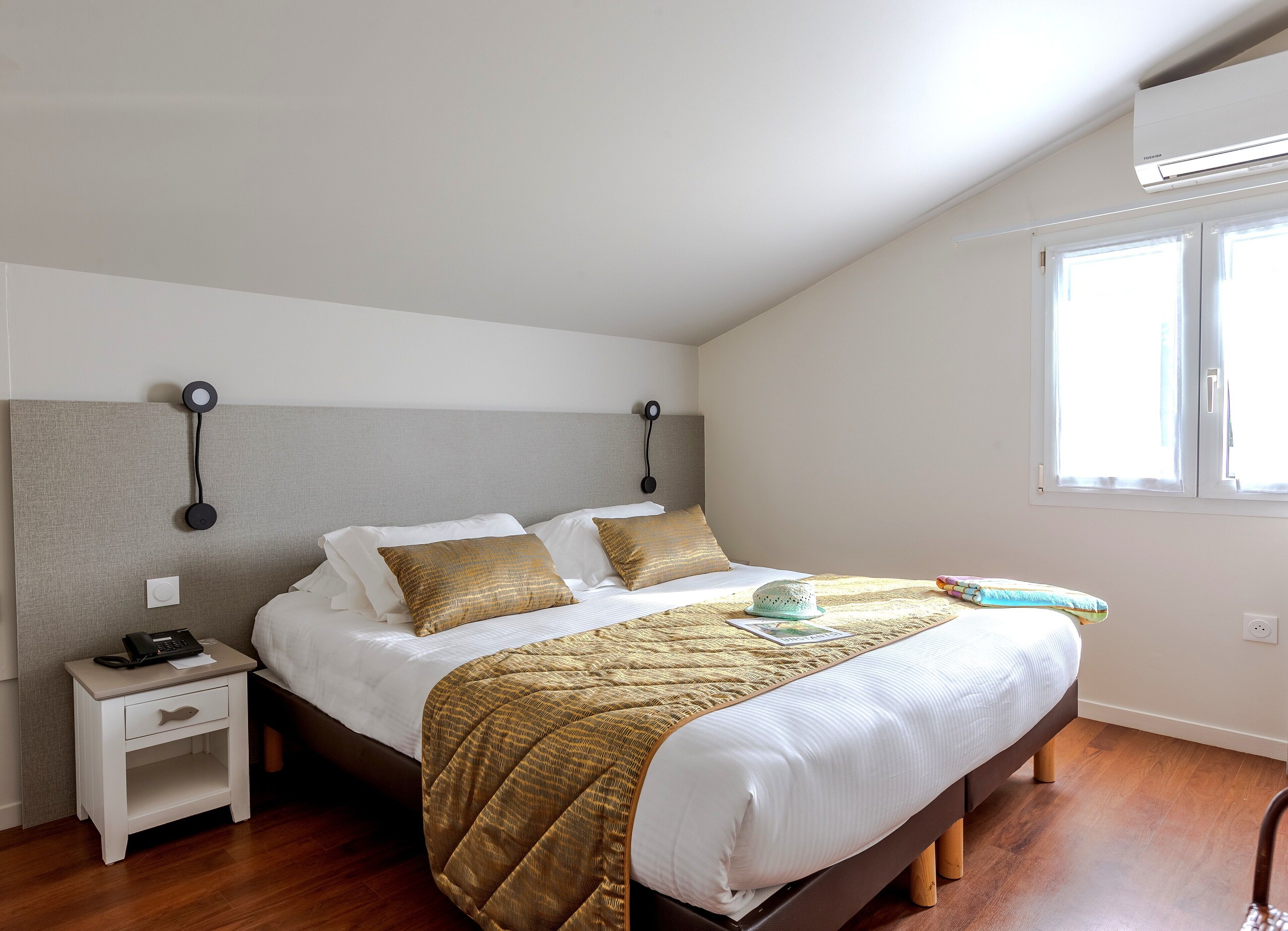 A neatly made bed with a gold quilt, a bedside table with a phone, and a window with curtains.