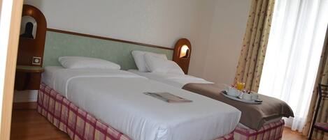 Une chambre d’hôtel avec deux lits, une tête de lit en bois, une table de chevet et une fenêtre avec des rideaux.