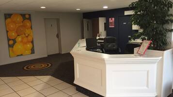 A reception area with a white reception desk, a large painting of oranges, a potted plant, and a carpeted floor.