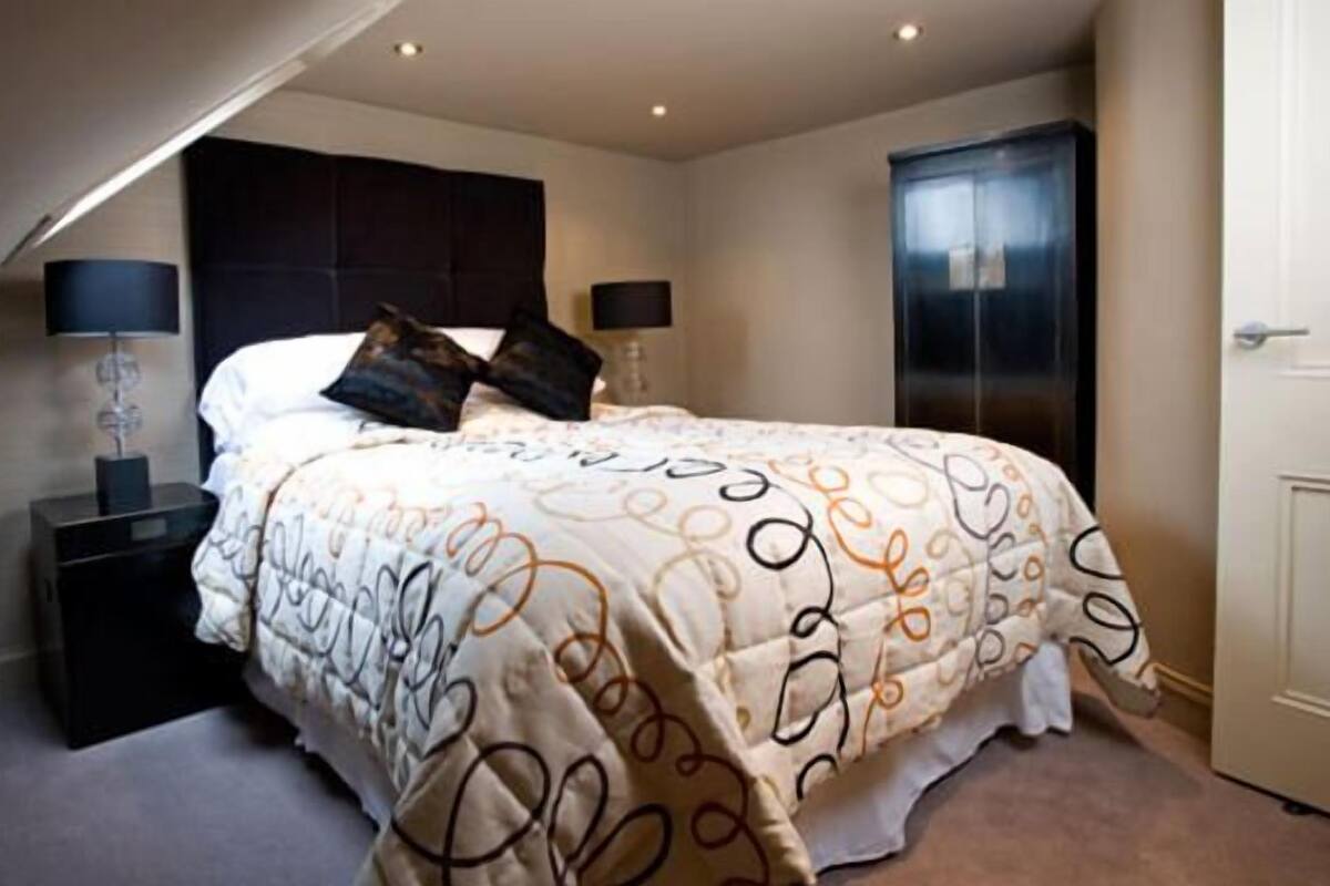 Quarto com cama grande, duas mesas de cabeceira com luminárias, cabeceira escura e um guarda-roupa.