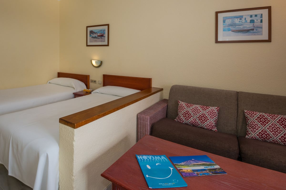 Habitación de hotel con dos camas individuales, un sofá, una mesa con un libro y una lámpara de pared.